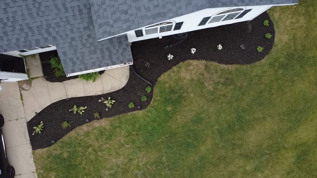 Aerial view of a landscaped yard featuring a curved flowerbed and plants near a white house.