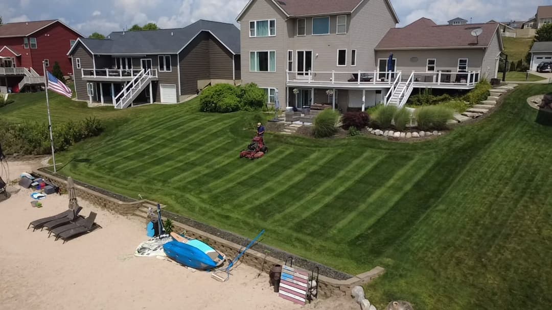 Lawn mowing in progress near waterfront homes with a beach and boats on display.