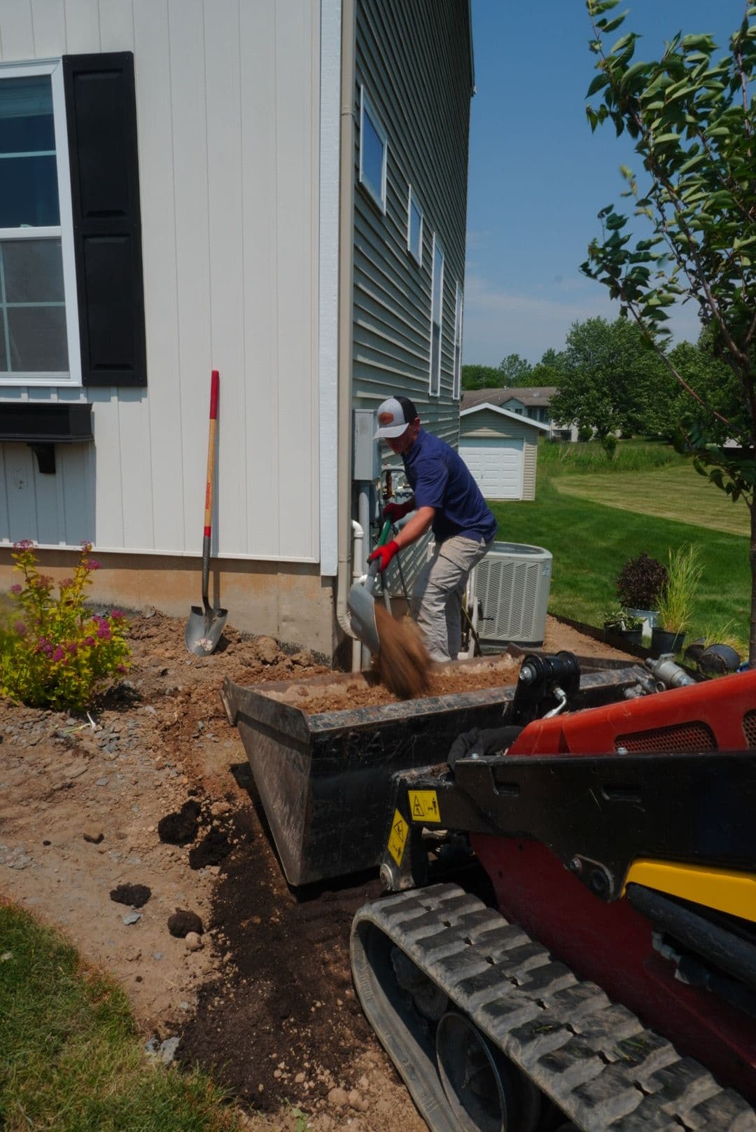 Man using a shovel to move dirt near a house with landscaping and construction equipment.