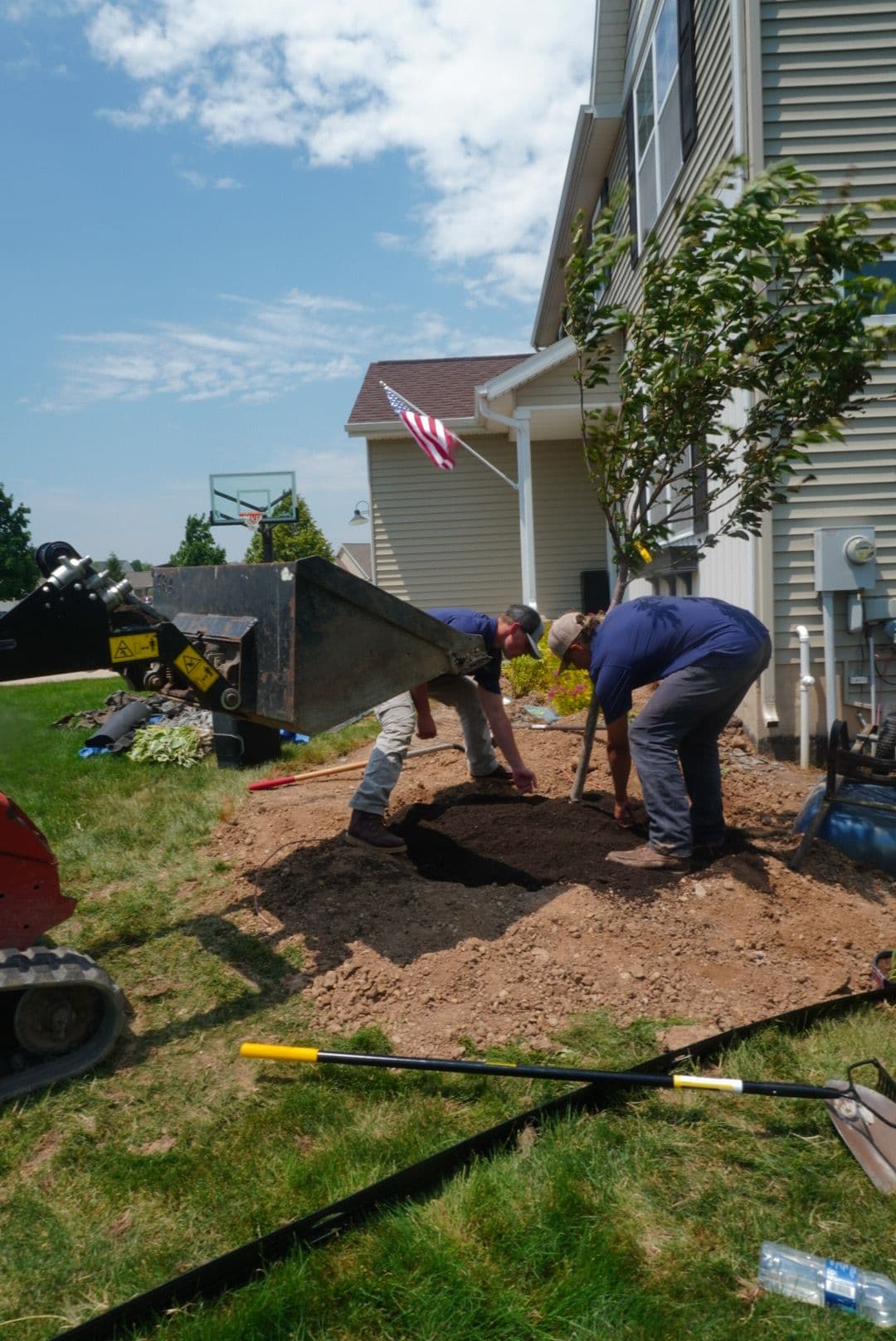 Workers digging a hole in a yard near a house with an American flag in the background.