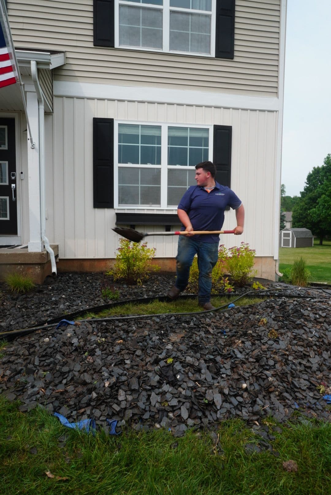 Man shoveling landscaping rocks in front of a house with an American flag.