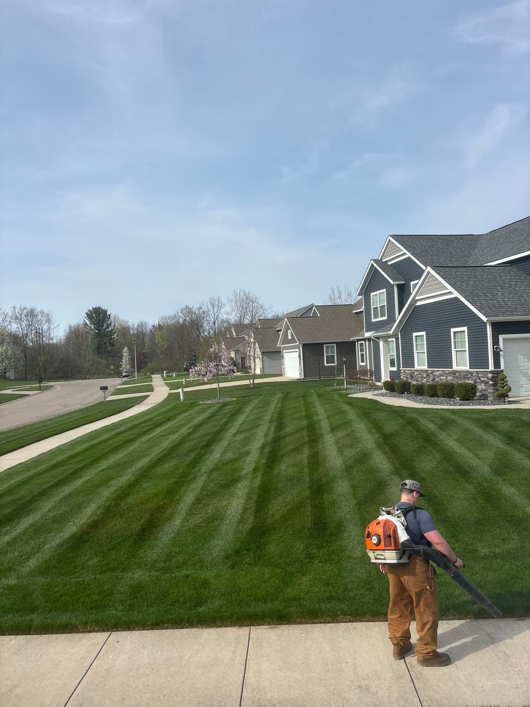Man using a leaf blower on a neatly striped lawn in a suburban neighborhood on a sunny day.