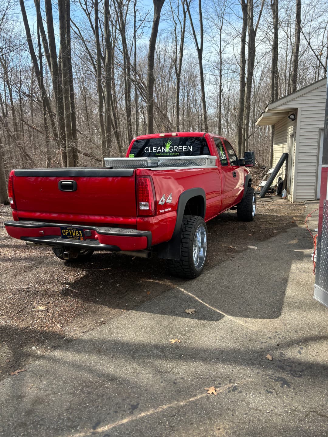 Red pickup truck with large tires parked near a house in a wooded area.