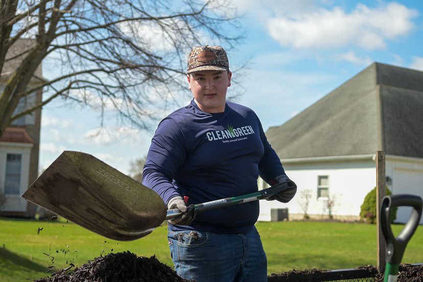Young man shoveling mulch in a yard, wearing a Clean Green shirt and gloves on a sunny day.